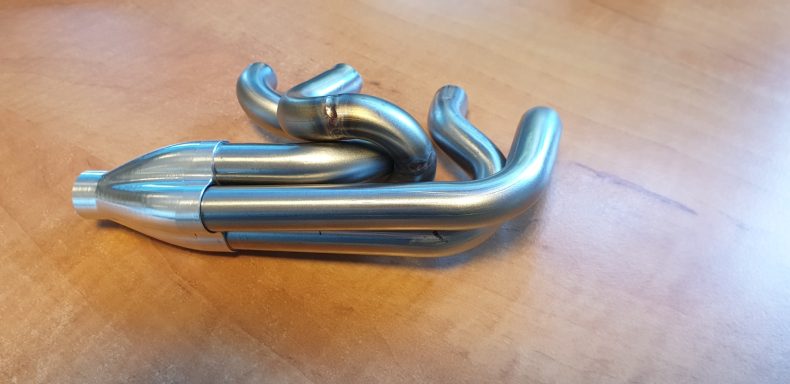 Uitlaatspruitstuk op schaal. Een geglazuurde metalen pijpstructuur met meerdere bochten en uiteinden.